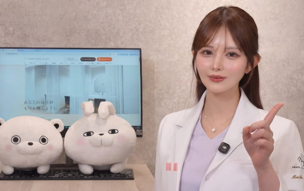 白衣の女性とPC画面とキャラクターのぬいぐるみ